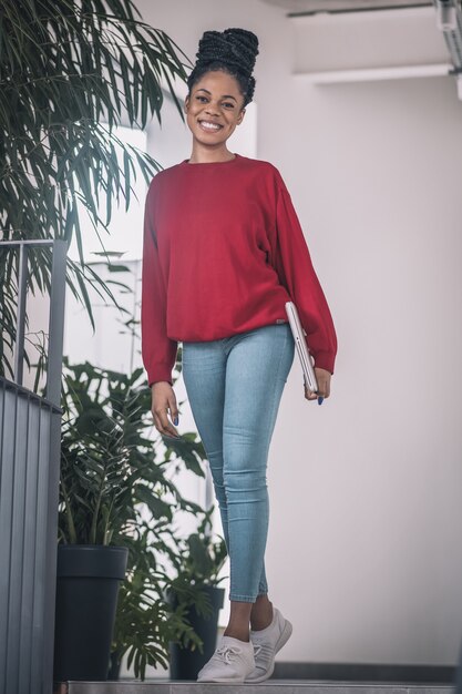 Dans le bureau. Femme noire en chemise rouge avec un ordinateur portable souriant et marchant dans le bureau