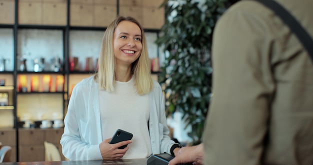 Dans le café, un barista prépare du café à emporter pour une cliente qui paie par téléphone portable sans contact au système de carte de crédit.