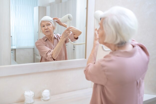 Dans le centre de spa. Femme mature aux cheveux gris séchant ses cheveux dans un vestiaire au centre de spa