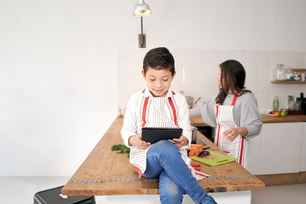 Dans la cuisine un enfant joue à des jeux vidéo avec une tablette pendant que sa mère coupe des légumes pour le repas...