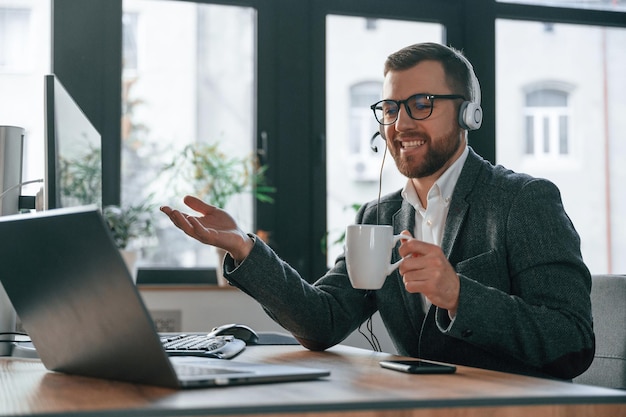 Dans des écouteurs ayant une conversation en ligne homme d'affaires en costume est dans le bureau avec l'ordinateur