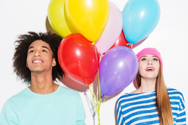 Photo dans leur propre monde coloré. funky jeune couple tenant des ballons et levant les yeux en se tenant debout sur fond blanc