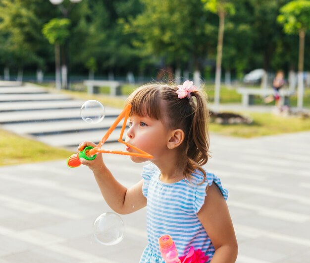 Dans un parc public, une petite fille souffle et attrape des bulles de savon. L'enfant joue et rit. Les bulles scintillent au soleil et volent dans différentes directions.