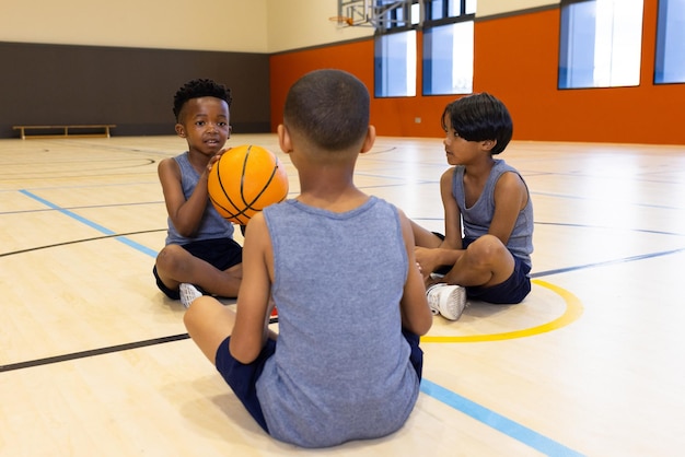 Dans la salle de sport de l'école, des garçons multiraciaux assis sur le sol tenant un ballon de basket-ball en train de parler ensemble.