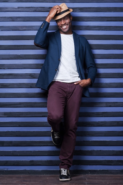 Photo dans son propre style. toute la longueur du beau jeune homme africain ajustant son chapeau et souriant en se tenant debout sur fond rayé