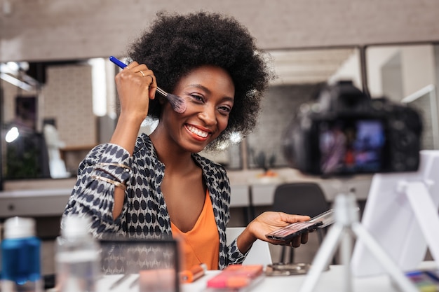 Dans le studio de mode. Femme positive à la peau foncée aux cheveux bouclés donnant une leçon vidéo en position assise