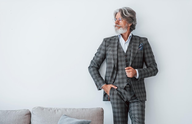 Dans des vêtements élégants Debout contre un mur blanc Senior homme moderne et élégant aux cheveux gris et à la barbe à l'intérieur