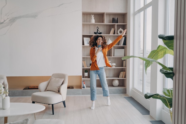 Danse énergique fille tenant un smartphone écoutant de la musique par des applications mobiles dans un salon moderne