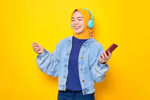 Danse jeune femme asiatique en veste de jeans portant des écouteurs pour écouter de la musique via un téléphone portable isolé sur fond jaune