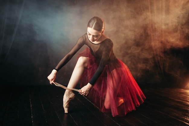 Danseuse de ballet en robe rouge assis sur le sol de la scène dans le théâtre et attachant des pointes. Formation de ballerine élégance en classe
