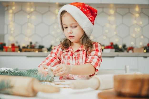 Dark-haired girl 3 ans in red Christmas cap et chemise à carreaux découpe des biscuits de pain d'épice de pâte roulée