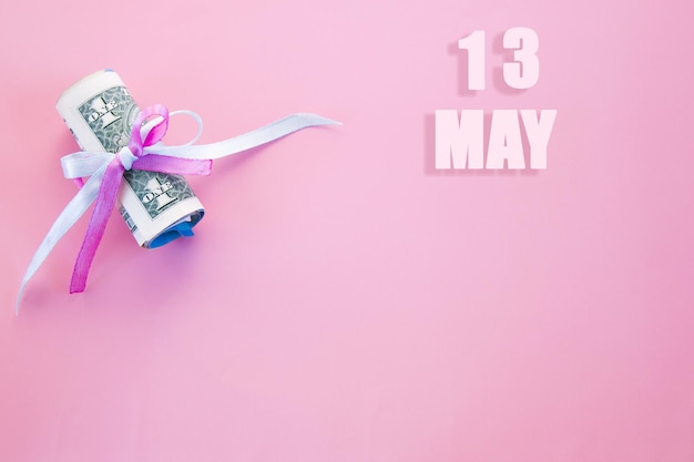 Date du calendrier sur fond rose avec des billets d'un dollar enroulés épinglés par un ruban rose et bleu le 13 mai