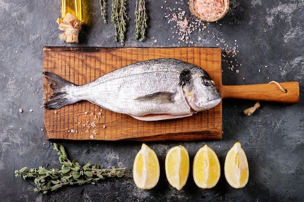 Daurade fraîche éviscérée crue ou poisson dorado servi avec citron, salade verte, romarin, huile d'olive et sel rose sur une planche à découper en bois sur une surface de texture sombre. Mise à plat, copiez l'espace.