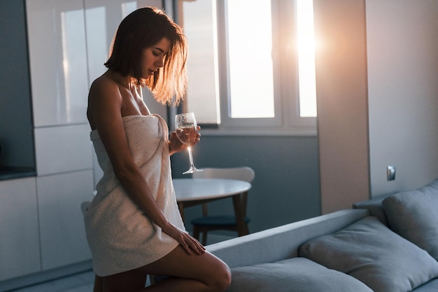 Debout au lever du soleil dans une serviette blanche Jeune femme est à l'intérieur dans la chambre d'une maison moderne pendant la journée