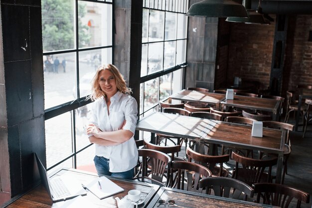 Debout les bras croisés. Femme d'affaires aux cheveux blonds bouclés à l'intérieur au café pendant la journée.