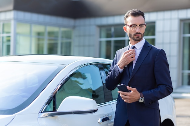 Debout près de la voiture. Bel homme d'affaires aux cheveux noirs prospère portant des lunettes debout près de sa voiture