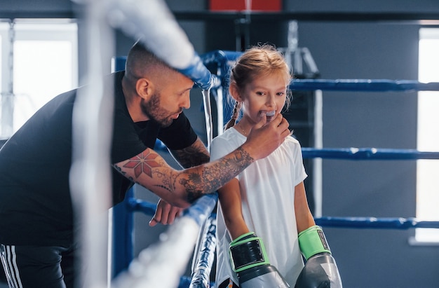 Photo debout sur le ring de boxe. le jeune entraîneur de boxe tatoué enseigne la petite fille mignonne dans le gymnase.