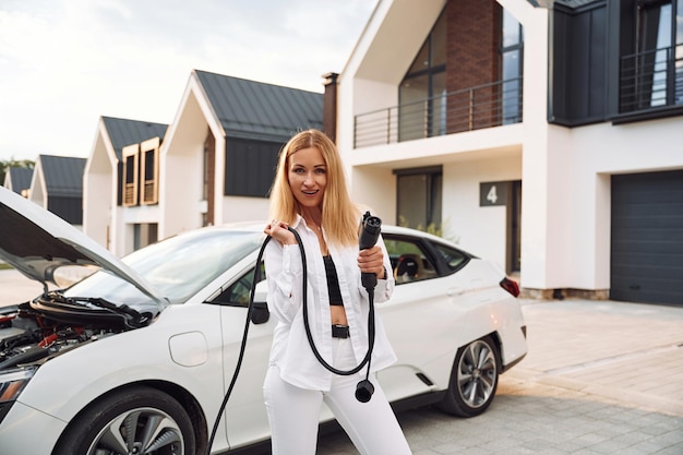 Debout et tenant le câble de charge Jeune femme en vêtements blancs est avec sa voiture électrique pendant la journée