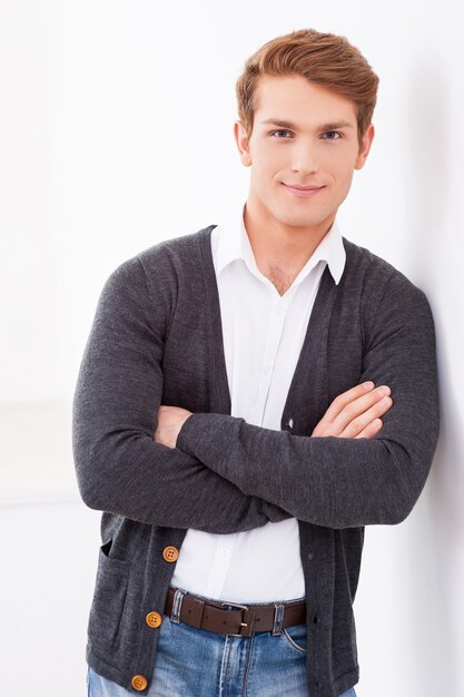 Décontracté beau. Beau jeune homme gardant les bras croisés et souriant à la caméra tout en se penchant contre le mur