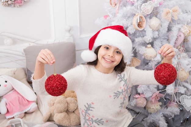 Décor de fantaisie. Créer une ambiance festive. Enfant décorant le sapin de Noël avec une boule rouge. Enfant de fille décorant l'arbre de Noël. Activité de vacances chérie. Enfant en bonnet de Noel décorant l'arbre de Noël.