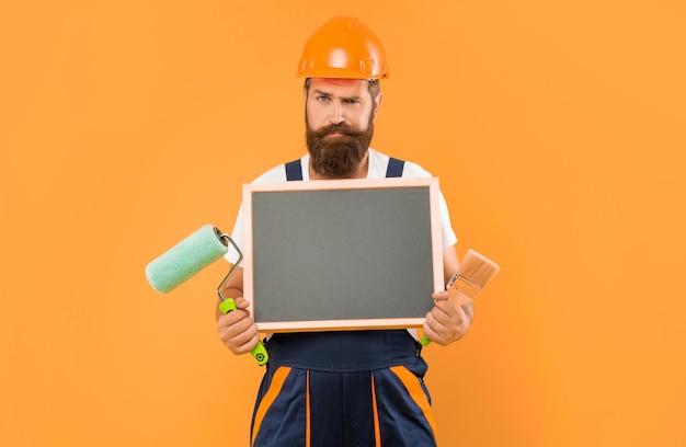 Décorateur d'homme sérieux en casque et vêtements de travail tenir pinceau rouleau à peinture et tableau noir