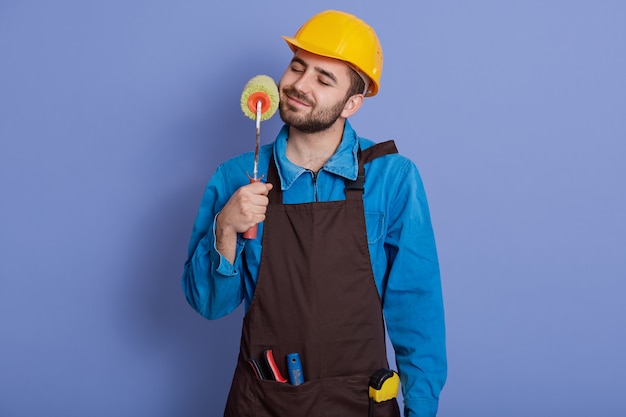 Décorateur avec rouleau à peinture dans les mains touchant sa barbe et garde les yeux fermés, gars portant un casque et un tablier marron