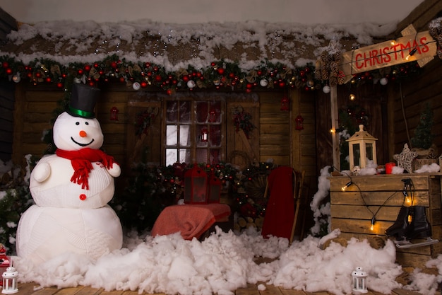 Décoration de Noël attrayante à l'intérieur d'une maison en bois avec un grand bonhomme de neige d'hiver, de la neige de coton sur le sol et un stand de Noël en bois éclairé.