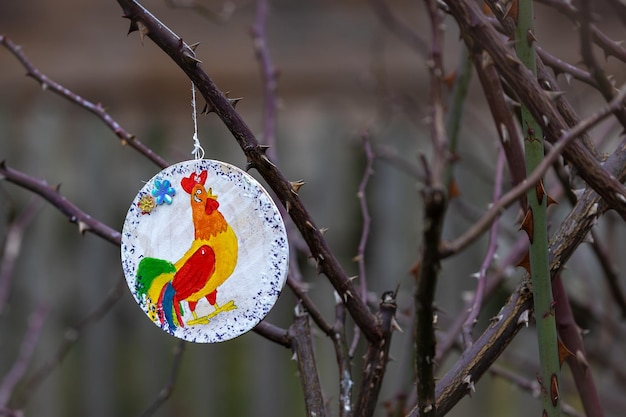 Une décoration de Noël faite à la main est accrochée à un arbre à l'extérieur. Un coq est dessiné sur le décor