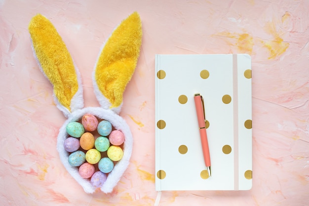 Décoration d'oeufs de couleur pastel, oreilles de lapin, carnet et stylo sur l'espace de travail de bureau, fond rose. Concept de célébration de Pâques. Vue de dessus, mise à plat, espace copie