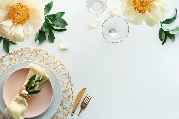 Décoration de table d'anniversaire dorée sans effort. Fleurs de pivoine jaune pâle et mise à plat de la fin du printemps et de l'été. Table à dîner blanche, ustensiles blancs et dorés, décorés de fleurs, lieu de texte