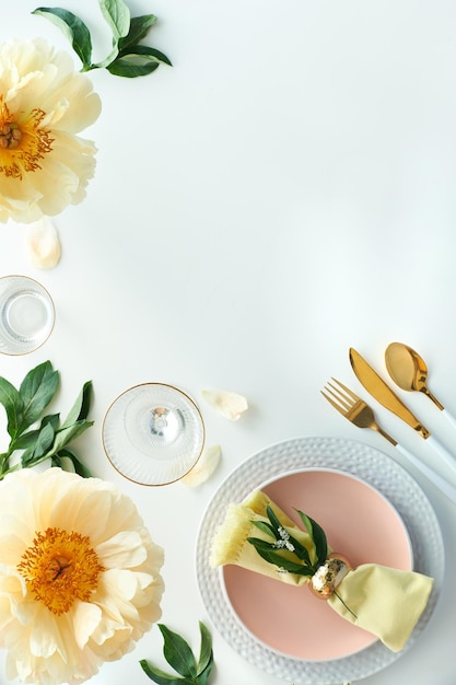 Décoration de table d'anniversaire dorée sans effort. Fleurs de pivoine jaune pâle et mise à plat de la fin du printemps et de l'été. Table à dîner blanche, ustensiles blancs et or, décorée de fleurs, espace de copie.