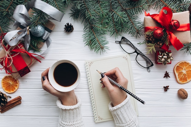 Décorations de Noël, cadeaux et nourriture sur un fond en bois blanc. Femme buvant un café et écrit pour faire la liste sur un cahier vintage.