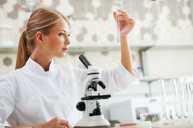 Découvrir la guérison. Jeune femme scientifique concentrée tenant un tube à essai et le regardant alors qu'elle était assise sur son lieu de travail