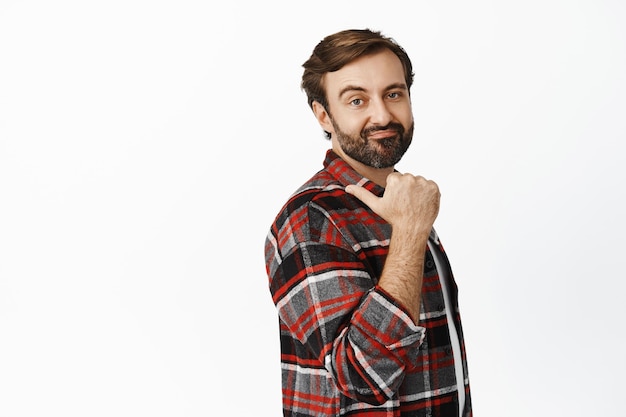 Déçu barbu en chemise à carreaux grimaçant et pointant vers la gauche montrant quelque chose avec aversion debout sur fond blanc