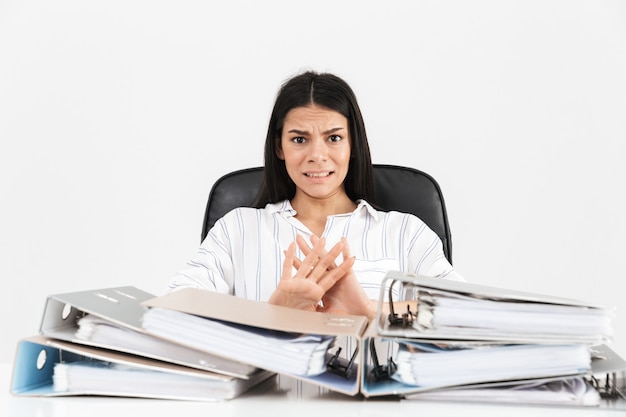 Déçu brunette businesswoman travaillant et assis au bureau avec tas de dossiers papier sur table isolé sur mur blanc