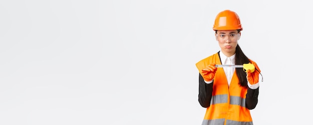 Déçu sombre femme asiatique ingénieur en construction architecte montrant une petite taille sur un ruban à mesurer faisant la moue contrarié par des mesures debout mécontent dans un casque de sécurité fond blanc