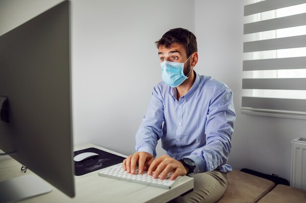 Dédié jeune homme d'affaires attrayant avec masque de protection assis au bureau et rapport de frappe.