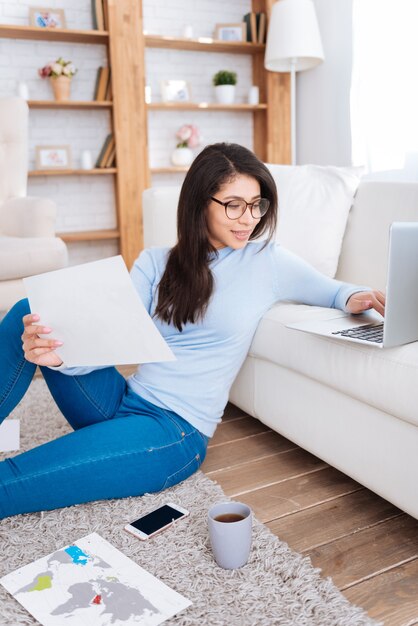 Défi éducatif. Charmante étudiante attrayante assise sur le sol tout en utilisant un ordinateur portable et en étudiant des documents