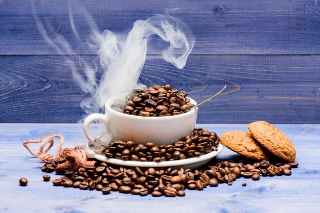 Degré de torréfaction du grain. Carte des boissons du café. Grains de café fraîchement torréfiés. Café pour l'inspiration et la charge énergétique. Tasse à café pleine de grains torréfiés bruns nuages blancs de fond en bois bleu fumée.