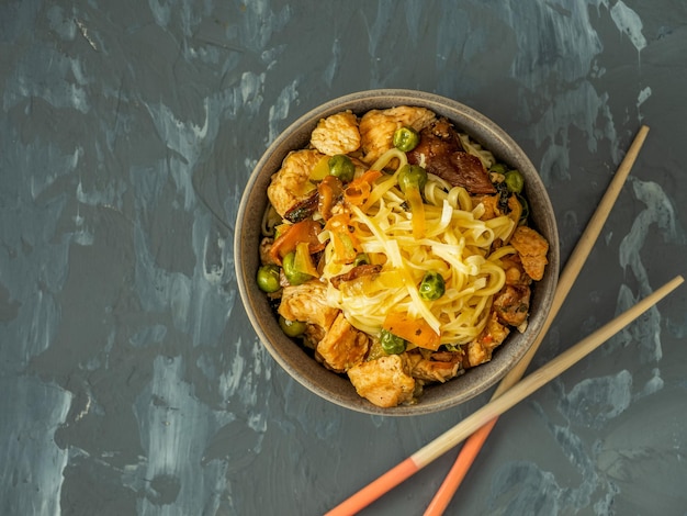 Déjeuner avec des nouilles, de la viande de dinde et des légumes dans une tasse en céramique profonde et des bâtons en bois