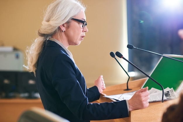 Délégué féminin élégant mature en costume parlant au microphone en se tenant debout par la tribune dans la salle de conférence au sommet