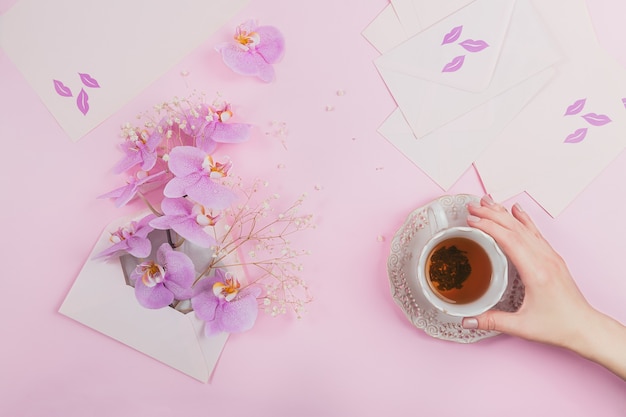 Délicate composition flatlay avec tasse de thé du matin, sac de lettre rose plein de fleurs d'orchidées pourpres et enveloppe vide sur une surface rose clair