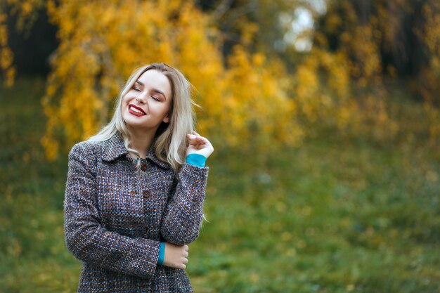 Délicieuse femme blonde aux lèvres rouges portant un manteau élégant profitant du temps dans la forêt d'automne. Espace libre