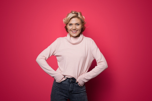 Délicieuse femme de race blanche aux cheveux blonds, souriant à la caméra dans un pull rose sur un mur rouge