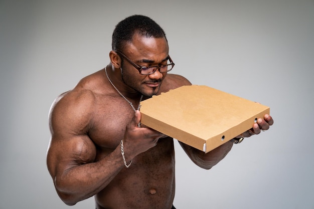 Délicieuse pizza Homme afro-américain appréciant une pizza savoureuse Homme tenant une boîte de pizzeria debout sur fond de studio gris avec les yeux fermés Cheat repas nutrition et restauration rapide suralimentation