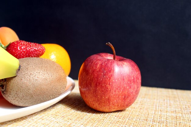 Une délicieuse pomme mûre se trouve sur la table à côté d'une assiette avec des fruits et des baies sur la table, sur un mur noir. Lumière directe dure.