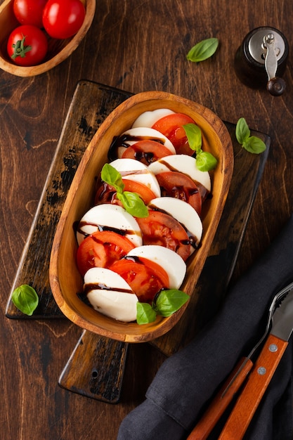 Photo délicieuse salade caprese italienne avec tomates mûres, basilic frais et fromage mozzarella dans un bol en bois d'olive sur la vieille table rustique vue de dessus à plat