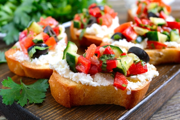 Délicieuses bruschettas au chèvre et légumes frais. Tranches de baguette croustillantes avec feta, tomates, concombres, poivrons, olives, herbes. Petit-déjeuner sain.