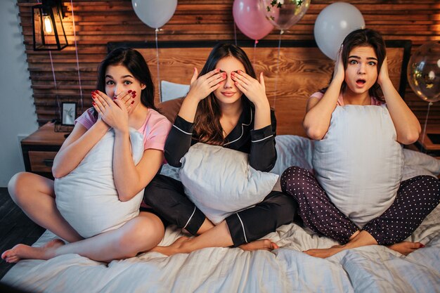 Photo délicieuses et drôles de jeunes femmes s'assoient sur le lit dans une salle de fête. ils gardent les jambes croisées. les premières femmes se couvrent la bouche, une seconde le fait avec les yeux. la troisième fille garde les oreilles fermées.