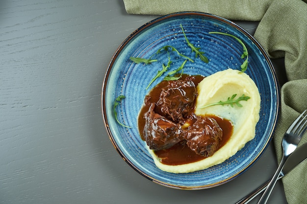 Délicieuses joues de veau avec sauce au miel et garniture en purée dans une assiette bleue sur une table en bois. Vue de dessus avec espace copie